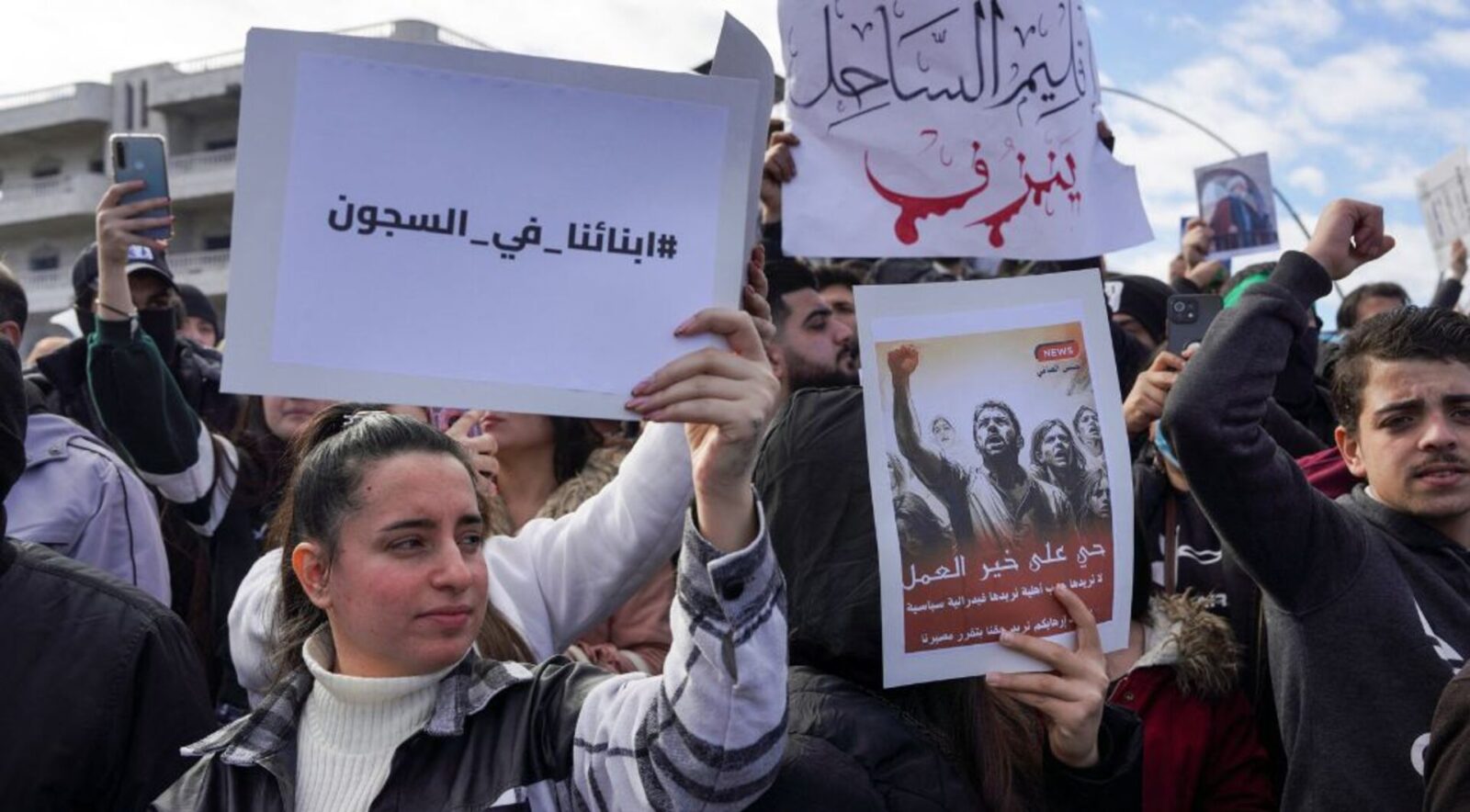 متظاهرون سوريون في مدينة ساحلية، مع خلفية تظهر قوات الأمن، تعكس التوترات الطائفية المتصاعدة.