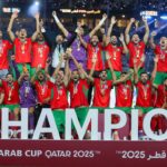 ** صورة لمدرب المنتخب المغربي محمد وهبي يرتدي قميصًا يظهر عليه شعار المنتخب المغربي، مع خلفية تظهر عليها شعار كأس العرب.